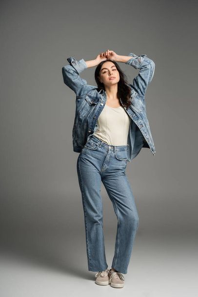 full length of chic woman with brunette hair posing with hands above head and closed eyes while standing in stylish blue jeans and denim jacket on grey background - Foto, imagen
