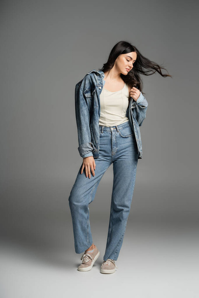 full length of charming woman with long brunette hair waving from wind posing in stylish blue jeans and denim jacket while standing on grey background - Foto, imagen