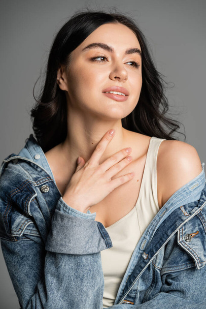 portrait of charming woman with brunette hair posing in stylish and blue denim jacket and holding hand near neck while looking away isolated on grey background - Photo, Image