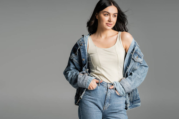 fashionable young woman with brunette hair standing with hands in pockets of blue jeans and posing in stylish denim jacket while smiling isolated on grey background - Foto, Imagem