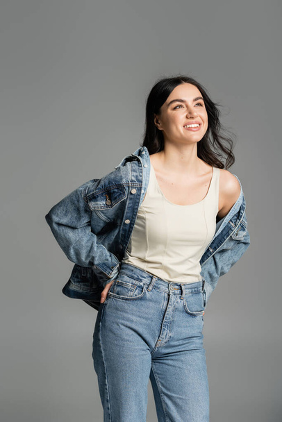 happy young woman with brunette hair standing in blue jeans and stylish denim jacket while smiling and posing with hand on hip isolated on grey background - 写真・画像