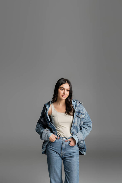 brunette young woman with gorgeous hair standing with hands in pockets of blue jeans and posing in stylish denim jacket while looking at camera isolated on grey background - Fotografie, Obrázek
