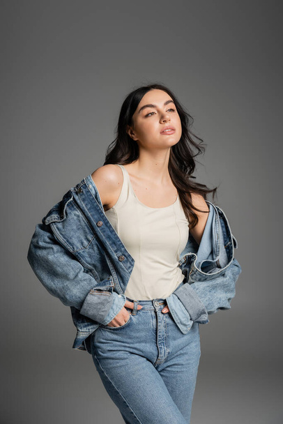 brunette young woman with shiny hair standing with hands in pockets of blue jeans and posing in stylish denim jacket while looking away isolated on grey background - Zdjęcie, obraz