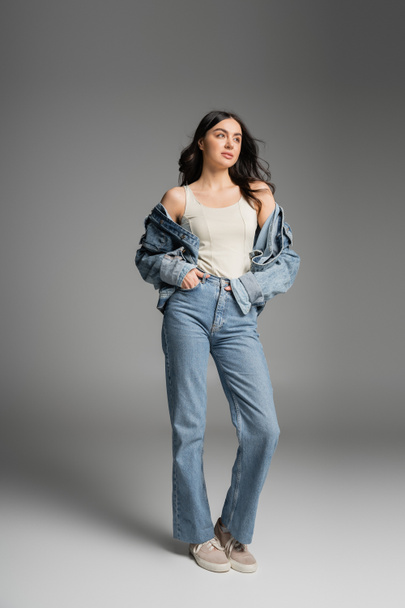 full length of young charming woman with long brunette hair posing in stylish blue jeans and denim jacket while standing with hands in pockets on grey background - Fotografie, Obrázek