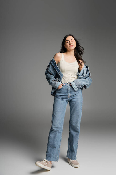 full length of young appealing woman with long brunette hair posing in stylish blue jeans and denim jacket while standing with hands in pockets on grey background - Zdjęcie, obraz