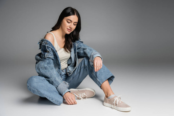 full length of young charming woman with long brunette hair and flawless natural makeup posing in stylish blue jeans and denim jacket while sitting on grey background - 写真・画像