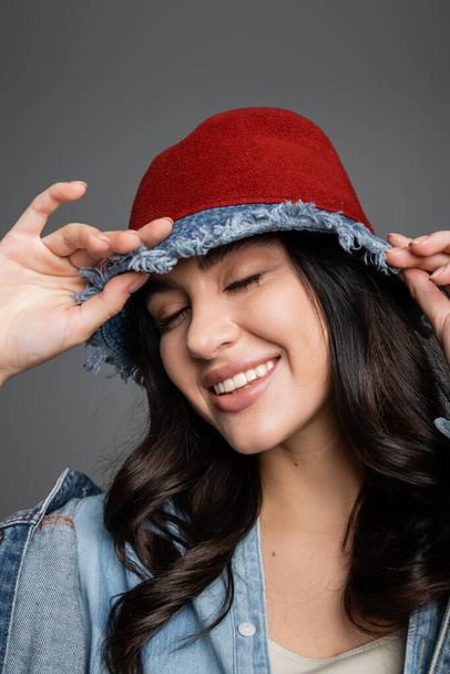 portrait of happy woman with closed eyes and flawless natural makeup posing in panama hat and denim jacket and smiling on grey background - Foto, afbeelding