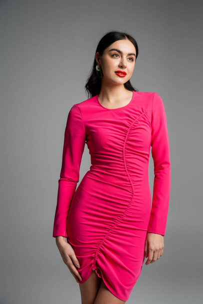 alluring young woman with shiny brunette hair, stylish earrings, red lips and trendy magenta dress looking away while posing on grey background  - Φωτογραφία, εικόνα