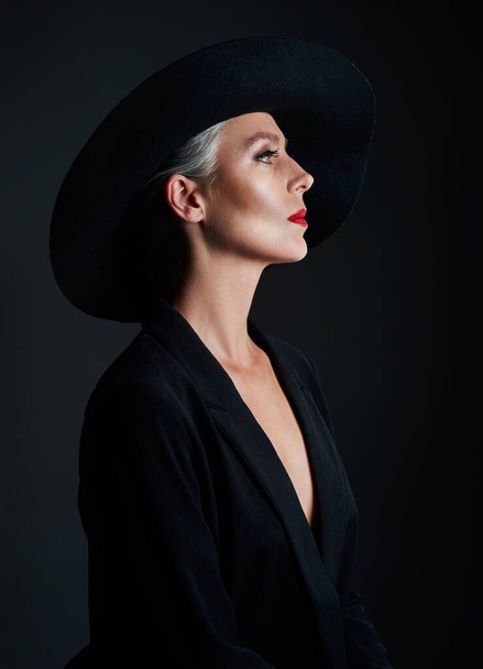 Ive never seen elegance go out of style. Studio shot of a beautiful mature woman wearing a hat and posing against a dark background - Foto, afbeelding