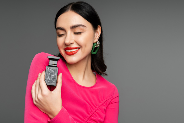 sophisticated young woman with shiny brunette hair, red lips, closed eyes and magenta dress holding bottle of perfume and smiling on grey background  - Foto, imagen