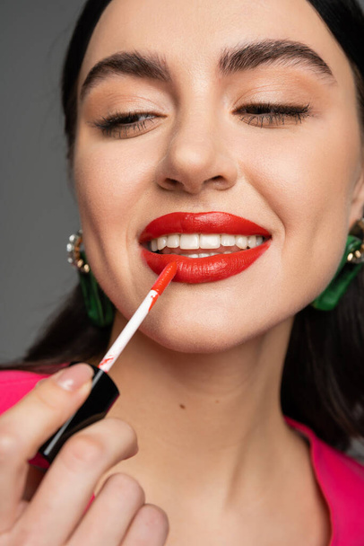 close up view of enchanting young woman with brunette hair and trendy earrings applying red lip gloss and smiling before party isolated on grey background  - Φωτογραφία, εικόνα