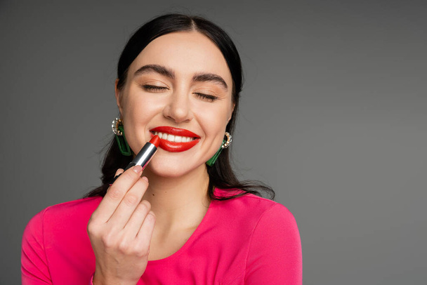 portrait of chic young woman with trendy earrings and flawless makeup applying red lipstick and smiling with closed eyes while posing on grey background  - Φωτογραφία, εικόνα