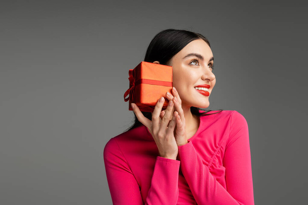 okouzlující a elegantní mladá žena s módní náušnice a lesklé brunetky vlasy s úsměvem při držení červené a zabalené dárkové krabice na šedém pozadí s kopírovacím prostorem  - Fotografie, Obrázek