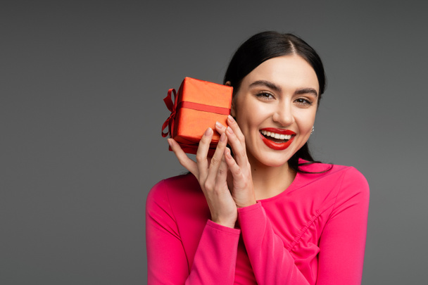 positive and chic young woman with trendy earrings and shiny brunette hair smiling while holding red and wrapped gift box on grey background  - Valokuva, kuva