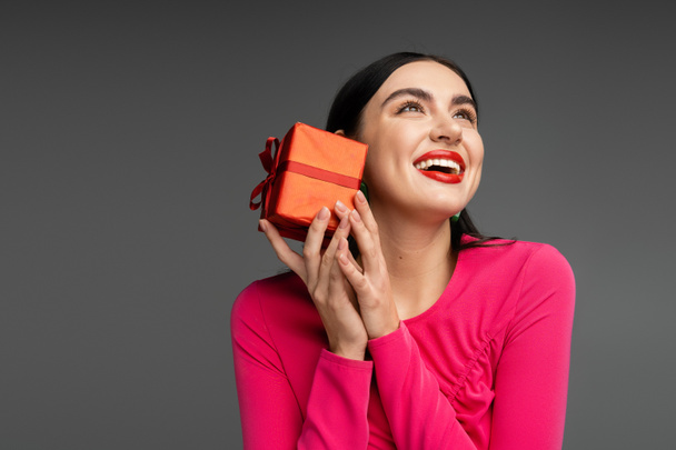 happy young woman with trendy earrings and shiny brunette hair smiling while holding red and wrapped gift box on holiday and looking up on grey background  - Foto, afbeelding
