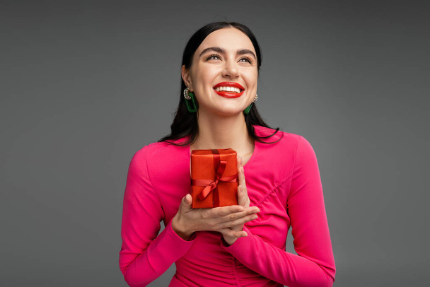 elegant and young woman with brunette hair and trendy earrings smiling while looking up and holding red and wrapped present for holiday on grey background  - Photo, Image