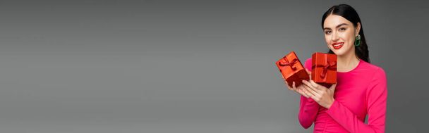 gelukkig vrouw met brunette haar glimlachen terwijl staan in magenta party jurk en het houden van verpakt en rode geschenkdozen voor vakantie op grijze achtergrond, banner  - Foto, afbeelding