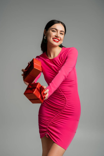 joyful and charming woman with brunette hair and trendy earrings smiling while standing in magenta party dress and holding wrapped gift boxes for holiday on grey background  - Foto, imagen