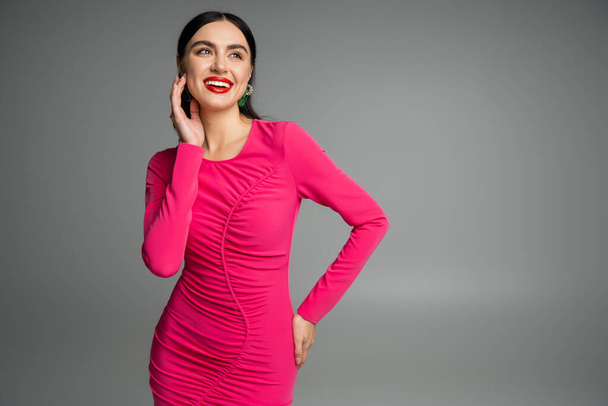elegant and alluring woman with brunette hair and trendy earrings standing in magenta  party dress while posing with hand on hip and smiling isolated on grey background  - Valokuva, kuva