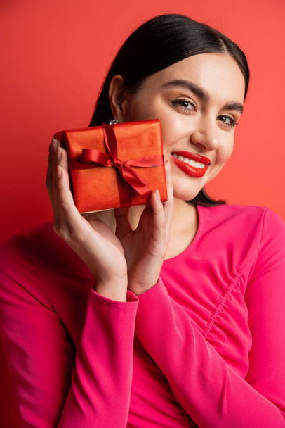 portrait of gorgeous woman with brunette hair smiling while standing in magenta party dress and holding wrapped surprise gift for holiday on red background  - Fotó, kép