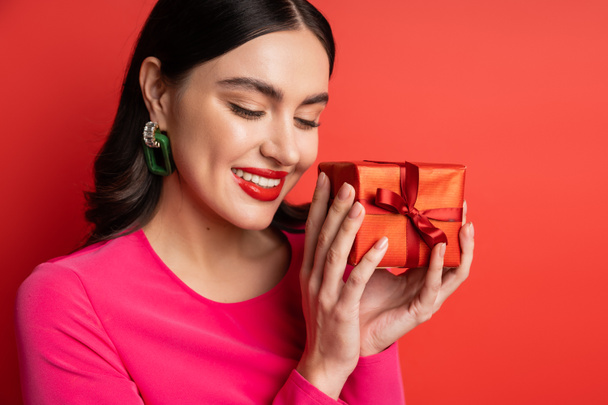 alluring woman with brunette hair and trendy earrings smiling while standing in magenta party dress and holding wrapped surprise gift for holiday on red background  - Foto, imagen