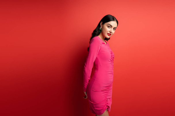glamorous woman with brunette hair and trendy earrings standing in magenta party dress while posing and looking at camera on red background - Photo, Image