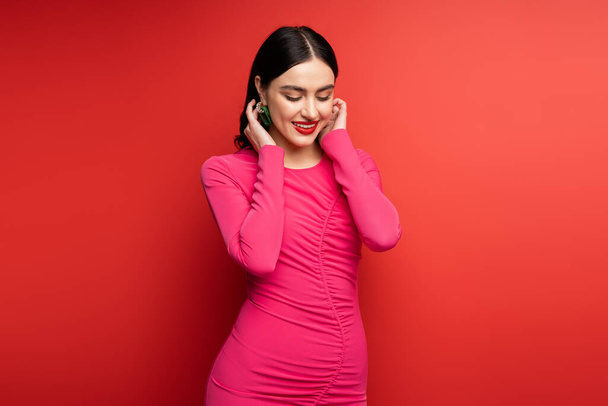 glamorous woman with brunette hair and trendy earrings smiling while standing in magenta party dress while posing and looking down on red background - Photo, Image
