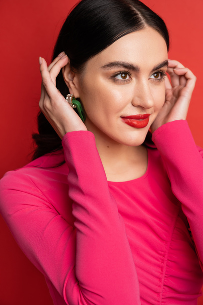 portrait of alluring woman with brunette hair and trendy earrings smiling while standing in magenta party dress while posing and looking away on red background - Foto, afbeelding