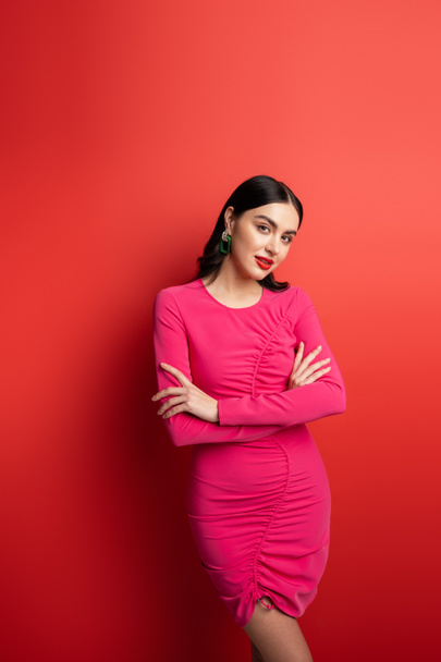gorgeous woman with brunette hair and trendy earrings standing with folded arms in magenta party dress while posing and looking at camera on red background - Photo, Image