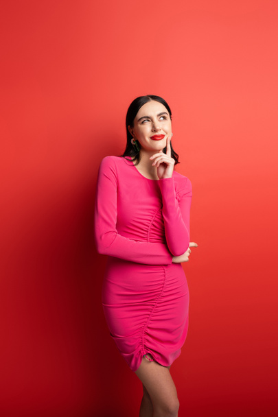 charming woman with brunette hair and trendy earrings smiling while thinking and holding hand near face and standing in magenta party dress on red background - Photo, image