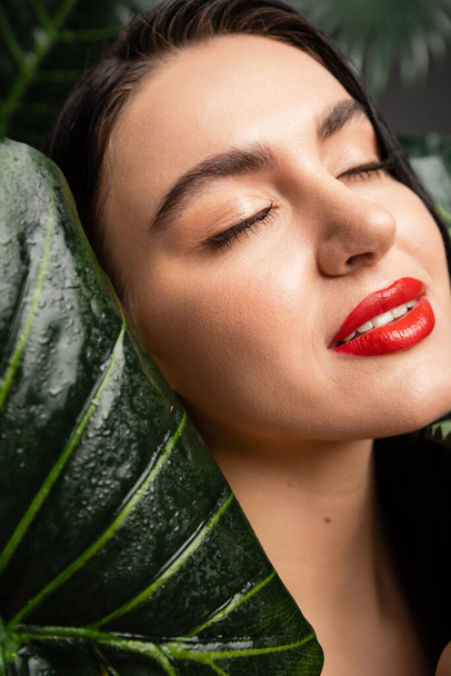 close up view of alluring young woman with brunette hair and red lips smiling while posing with closed eyes around tropical, wet and green palm leaves with raindrops  - Foto, imagen