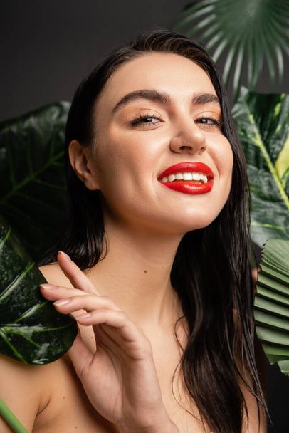 enchanting young woman with brunette hair and red lips smiling while posing around tropical, wet and green palm leaves with raindrops on them and looking at camera  - Foto, Bild
