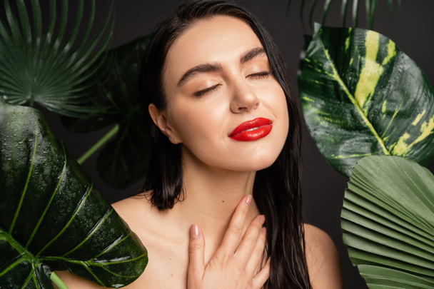gorgeous young woman with brunette hair and red lips smiling while posing with closed eyes around tropical, wet and green palm leaves with raindrops on them   - Fotografie, Obrázek