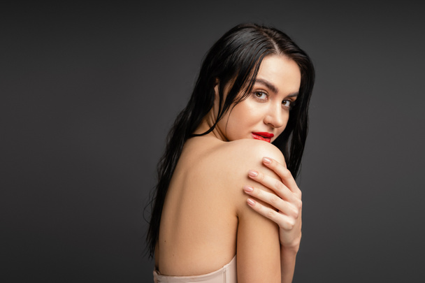 portrait of young and graceful woman with wet brunette hair and red lips touching bare shoulder while looking at camera after shower isolated on grey background  - Foto, imagen