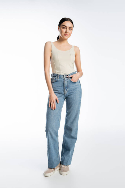 full length of charming young brunette woman standing in blue denim jeans and tank top while posing with hand in pocket and looking at camera on white background  - Φωτογραφία, εικόνα