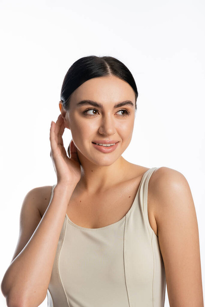 portrait of charming young woman with flawless and natural makeup standing in tank top while adjusting brunette hair and looking away isolated on white background  - Φωτογραφία, εικόνα