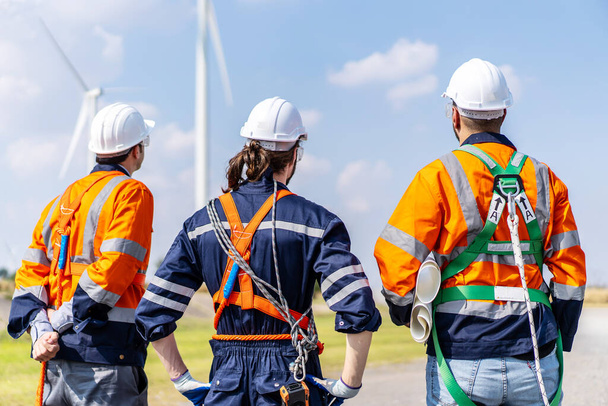 Rückansicht von drei kaukasischen Ingenieuren in Uniform und Hardhat sprechen über Systeminstallation auf landwirtschaftlichem Feld mit Windrädern, Windkraftanlagen erzeugen Strom, Erneuerbare Energien - Foto, Bild