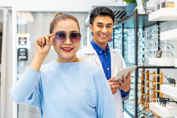 Mladý asijský oftalmolog představuje zákazníkovi novou kolekci brýlí, pomáhá a prodává seniorku v optickém centru, koncepci péče o oči, používá správnou asistenci nebo vadný zrak. - Fotografie, Obrázek