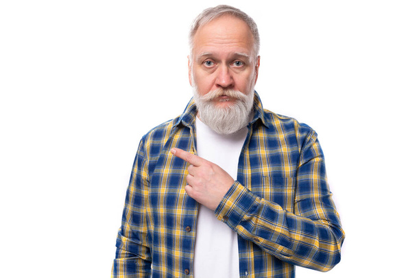 pensionista senior hombre de pelo gris con una barba en una camisa muestra su mano a un lado sobre un fondo blanco con espacio para copiar. - Foto, imagen