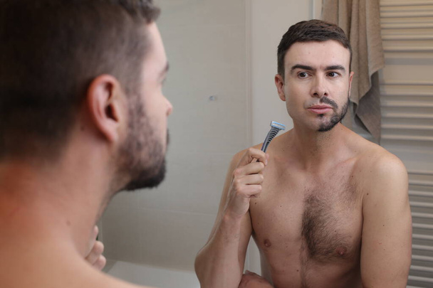 πορτρέτο του όμορφου νεαρού άνδρα με μισοξυρισμένο μούσι μπροστά από τον καθρέφτη στο μπάνιο - Φωτογραφία, εικόνα