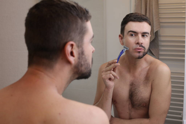 портрет красивого молодого человека с наполовину бритыми бородами перед зеркалом в ванной комнате - Фото, изображение