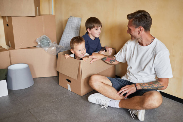 Aile yeni bir eve taşınırken iki oğluyla karton kutularda oynayan gülümseyen dövmeli baba portresi. - Fotoğraf, Görsel