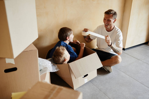 Retrato de alto ángulo del padre feliz jugando con dos hijos en caja de cartón durante la casa de mudanza familiar - Foto, imagen