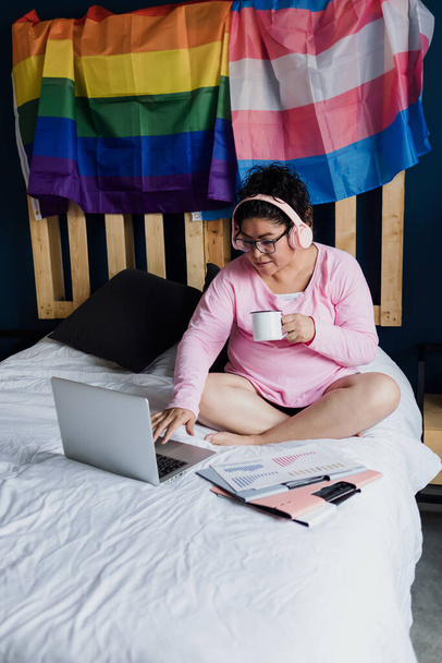 Латиноамериканська лесбіянка лежить на ліжку за допомогою ноутбука або комп "ютера, який навчається на прес - конференції вдома в Мексиці, іспаномовні гомосексуалісти з lgbt спільноти з райдужним прапором в Латинській Америці. - Фото, зображення
