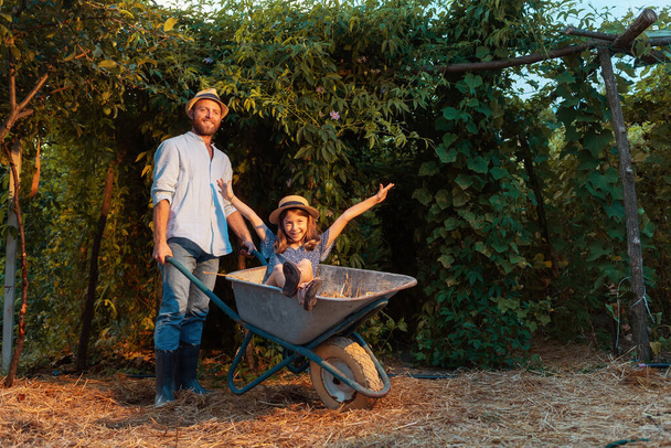 Smiling caucasian father in a straw hat holding a cart with a happy little daughter. Family weekends together in the backyard. The concept of gardening and farming. - Photo, image