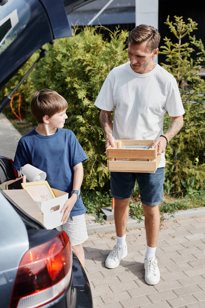 Vertikální portrét otce a syna nesoucích krabice k autu při stěhování do nového domu osvětleného slunečním světlem venku - Fotografie, Obrázek