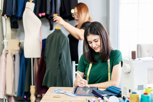 Кавказская модельер работает в студии, рисуя эскизы с цифровой планшет и красочные ткани для коллекции дизайна платья, выбирая цвета одежды для пошива и проектирования - Фото, изображение