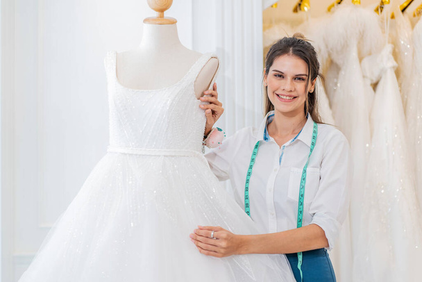 Lächelnde kaukasische Frau ist Brautmodenbesitzerin beim Aufräumen des Hochzeitskleides und blickt in die Kamera im Hochzeitsstudio, Kleinunternehmerin Hochzeitsplanerin und Maßdesignerin - Foto, Bild
