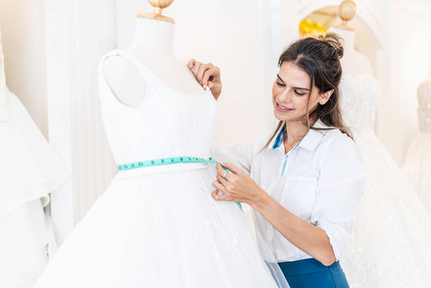 Lächelnde kaukasische Designerin oder Brautmodenbesitzerin mit Maßband misst Hochzeitskleid auf Schaufensterpuppe in Werkstatt, Kleinunternehmerin Hochzeitsplanerin und Maßdesignerin - Foto, Bild