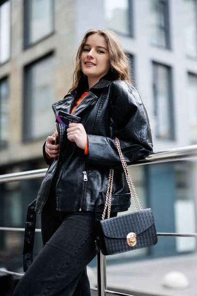 Young girl in leather jacket posed in urban city, holding credit card with handbag and phone. - Foto, Bild